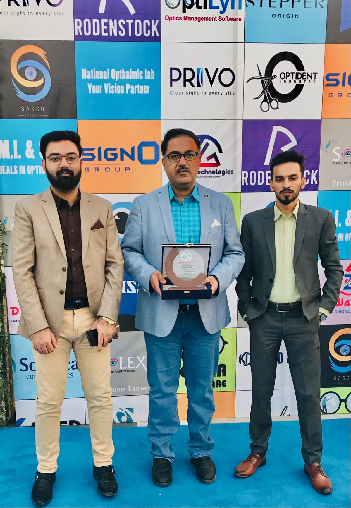 The appreciation award plaque presented to LitGrey, held open in front of the OptiPak 2025 sponsor wall
